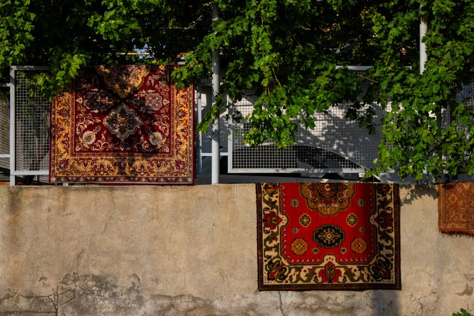 Close-up image of a rolled-up, intricately patterned Persian-style rug with floral and geometric designs in shades of red, blue, cream, and gold, positioned on a dark wooden floor. Bright natural ligh
