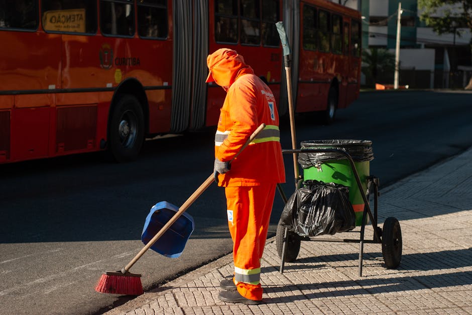 A worker dressed in high-visibility orange safety uniform, including a jacket and pants with reflective stripes, stands on an asphalt pavement, facing away from the camera. The worker is wearing a mat