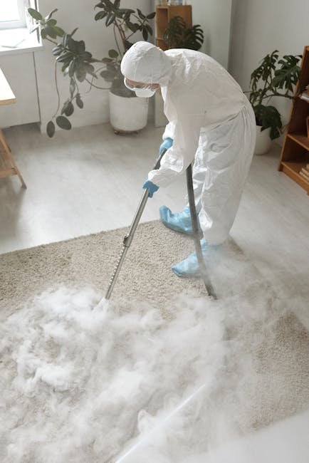A professional cleaner dressed in white protective clothing, including gloves and a face mask, is performing surface cleaning in a bright, modern living room. They are vacuuming a plush, light-colored