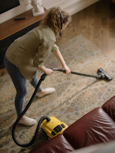A woman with curly hair, wearing a green shirt and blue jeans, is vacuuming a patterned area rug in a living room. She holds a yellow vacuum cleaner with a black hose, aiming the cleaner's nozzle at t