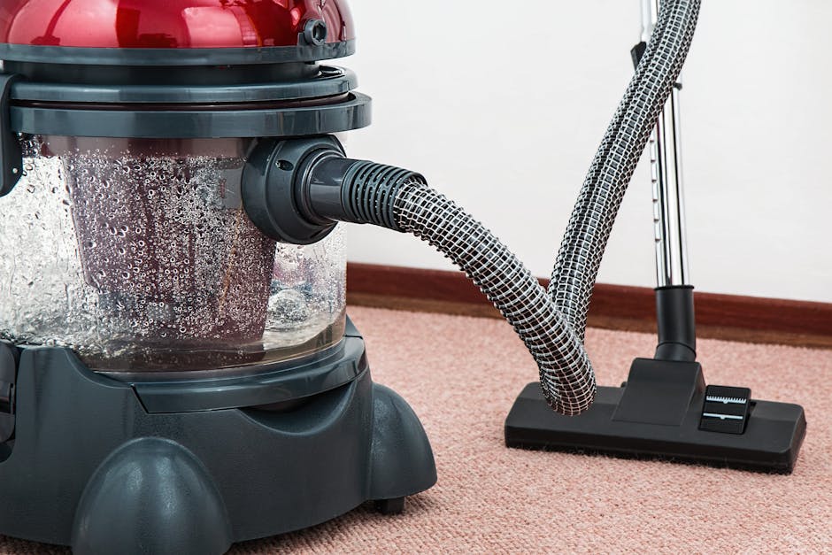 A woman with curly hair, wearing a green shirt and blue jeans, is vacuuming a patterned area rug in a living room. She holds a yellow vacuum cleaner with a black hose, aiming the cleaner's nozzle at t