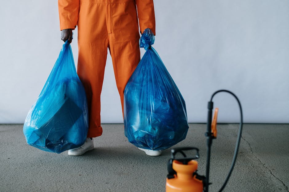 A person wearing orange work overalls and white shoes is standing on a light gray concrete floor, holding two large blue plastic trash bags filled with waste. The background is plain white, and part o