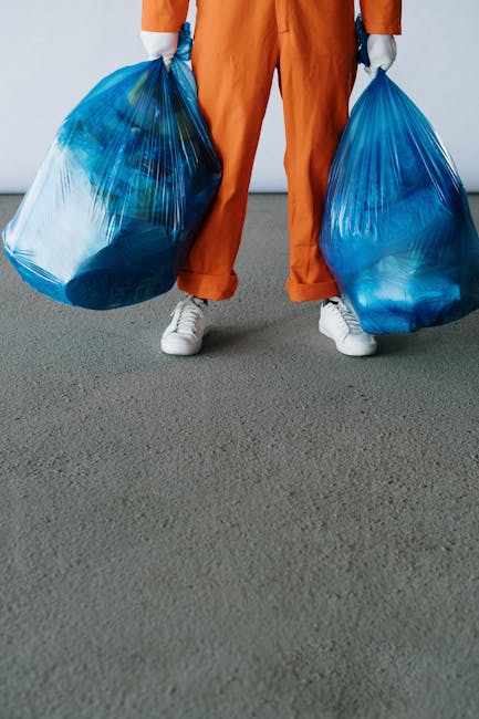 A person wearing orange work overalls and white shoes is standing on a light gray concrete floor, holding two large blue plastic trash bags filled with waste. The background is plain white, and part o