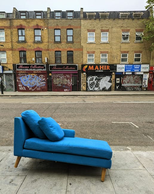A scenic view of a narrow canal in Islington, lined with lush green trees on both sides, with clear water reflecting the blue sky and scattered clouds above. Several small boats are moored along the c